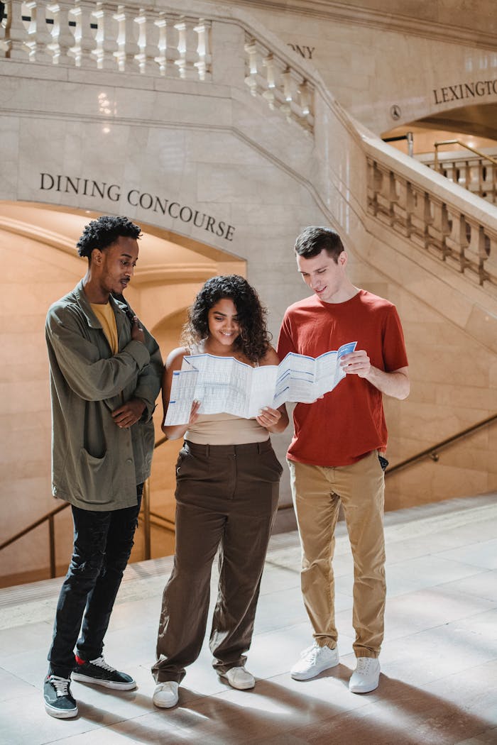 contact-img Full body of positive young multiracial friends reading map while standing near stairway in Grand Central Terminal during trip in New York